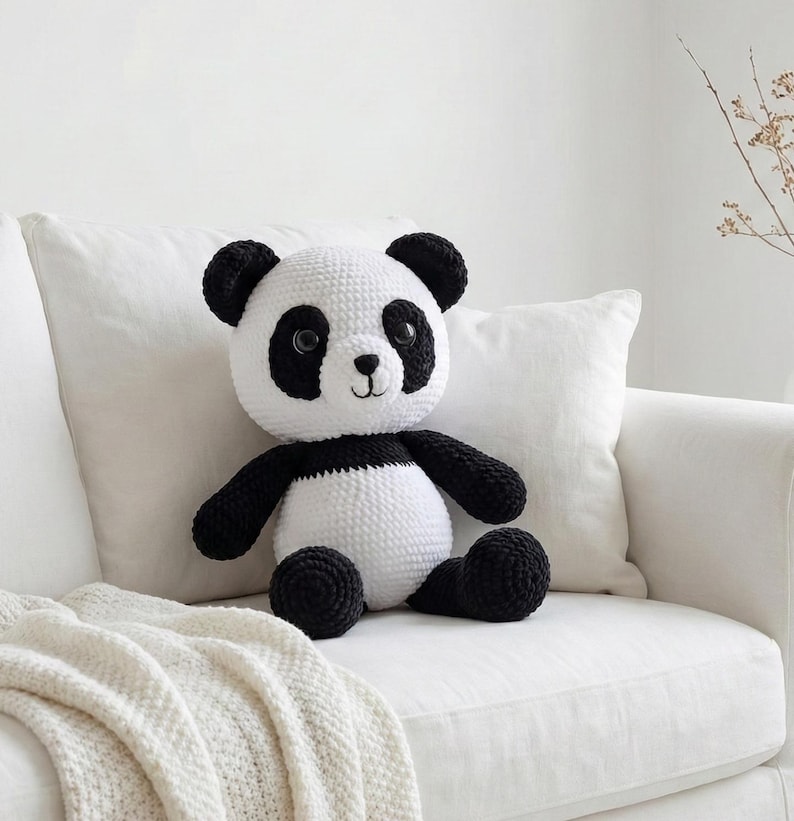 May include: A handmade crochet panda bear with a black and white color scheme. The panda has black eyes, ears, arms, and legs, with a white body and head. The stuffed animal is sitting on a white couch.