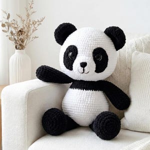 5 ALT TEXT FOR PHOTOS
1) Crochet panda plush sitting on a cozy chair with black ears, black eye patches, and a white face in a neutral room
2) Close-up of amigurumi panda face showing shiny eyes, black eye patches, and a small embroidered smile
3) Full view of crochet panda bear with round belly and chunky black arms and legs posed indoors
4) Side angle of crocheted panda plush highlighting the rounded head, ears, and smooth black-and-white stitch texture