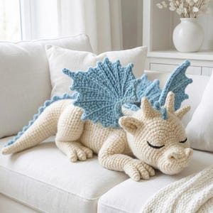 5 ALT TEXT FOR PHOTOS
1) Crochet sleeping dragon plush with pale blue wings resting on a white sofa in a bright neutral room
2) Close-up of the crocheted dragon head with embroidered sleepy eyes and soft textured stitches
3) Side view of amigurumi dragon showing sculpted wings, horns, and back spikes in pastel tones
4) Crochet dragon plush posed on a cozy knit blanket with minimalist nursery-style decor in the background