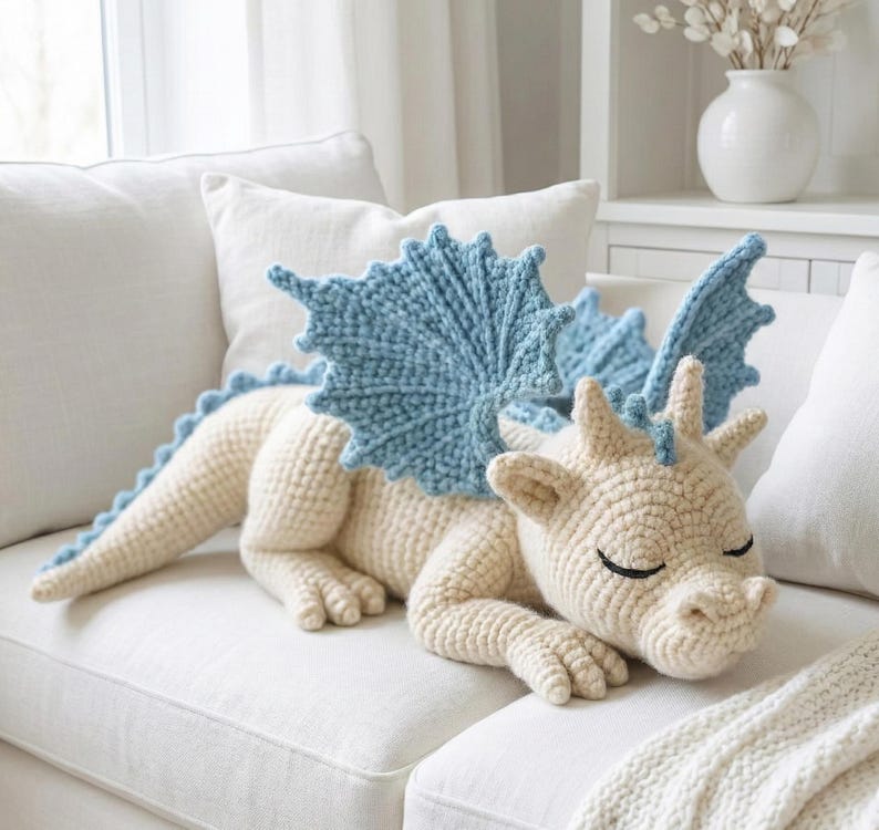 5 ALT TEXT FOR PHOTOS
1) Crochet sleeping dragon plush with pale blue wings resting on a white sofa in a bright neutral room
2) Close-up of the crocheted dragon head with embroidered sleepy eyes and soft textured stitches
3) Side view of amigurumi dragon showing sculpted wings, horns, and back spikes in pastel tones
4) Crochet dragon plush posed on a cozy knit blanket with minimalist nursery-style decor in the background