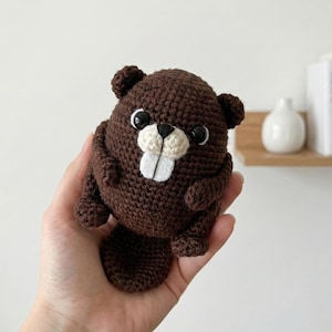 May include: A handmade, brown crochet beaver plush toy with black eyes and white teeth. The beaver has a rounded body, small arms, and a flat tail. The plush is held in a hand against a neutral background.