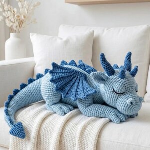 5 ALT TEXT FOR PHOTOS
1) Crochet sleeping dragon plush with pale blue wings resting on a white sofa in a bright neutral room
2) Close-up of the crocheted dragon head with embroidered sleepy eyes and soft textured stitches
3) Side view of amigurumi dragon showing sculpted wings, horns, and back spikes in pastel tones
4) Crochet dragon plush posed on a cozy knit blanket with minimalist nursery-style decor in the background
