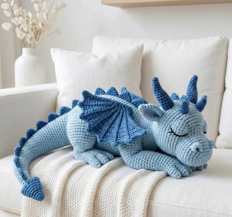 5 ALT TEXT FOR PHOTOS
1) Crochet sleeping dragon plush with pale blue wings resting on a white sofa in a bright neutral room
2) Close-up of the crocheted dragon head with embroidered sleepy eyes and soft textured stitches
3) Side view of amigurumi dragon showing sculpted wings, horns, and back spikes in pastel tones
4) Crochet dragon plush posed on a cozy knit blanket with minimalist nursery-style decor in the background