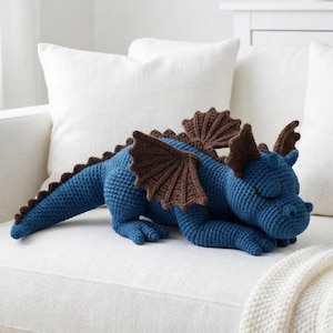 May include: A blue crocheted dragon with brown wings and accents rests on a white couch. The handmade plush toy features a long tail, spiky back, and closed eyes, suggesting a sleeping pose. A cozy, decorative item.