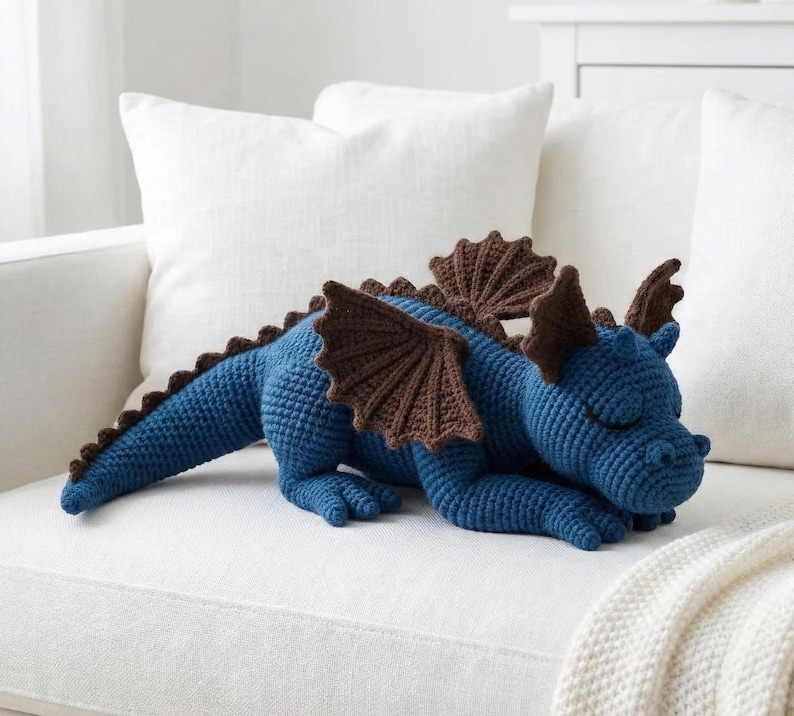 May include: A blue crocheted dragon with brown wings and accents rests on a white couch. The handmade plush toy features a long tail, spiky back, and closed eyes, suggesting a sleeping pose. A cozy, decorative item.