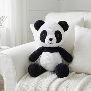 May include: A handmade crochet panda bear with black and white yarn. The panda has black eyes, ears, arms, and legs, with a white body. The stuffed animal is sitting on a white couch with white pillows and a white blanket.