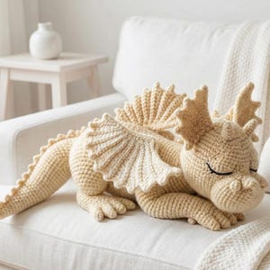 5 ALT TEXT FOR PHOTOS
1) Crochet sleeping dragon plush with pale blue wings resting on a white sofa in a bright neutral room
2) Close-up of the crocheted dragon head with embroidered sleepy eyes and soft textured stitches
3) Side view of amigurumi dragon showing sculpted wings, horns, and back spikes in pastel tones
4) Crochet dragon plush posed on a cozy knit blanket with minimalist nursery-style decor in the background