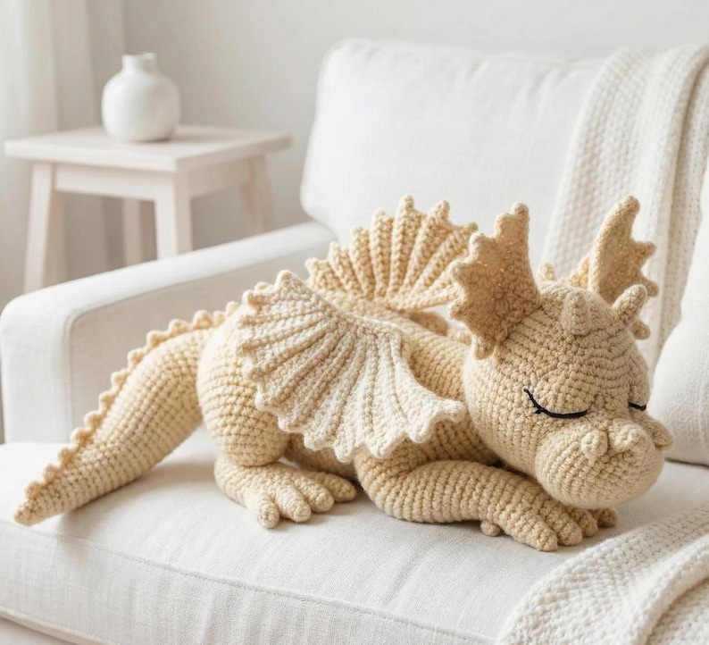 5 ALT TEXT FOR PHOTOS
1) Crochet sleeping dragon plush with pale blue wings resting on a white sofa in a bright neutral room
2) Close-up of the crocheted dragon head with embroidered sleepy eyes and soft textured stitches
3) Side view of amigurumi dragon showing sculpted wings, horns, and back spikes in pastel tones
4) Crochet dragon plush posed on a cozy knit blanket with minimalist nursery-style decor in the background
