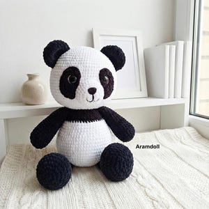 May include: A handmade crochet panda bear doll with a white body, black arms, legs, and ears. The panda has black eye patches and a small, embroidered smile. The doll is sitting on a white surface.