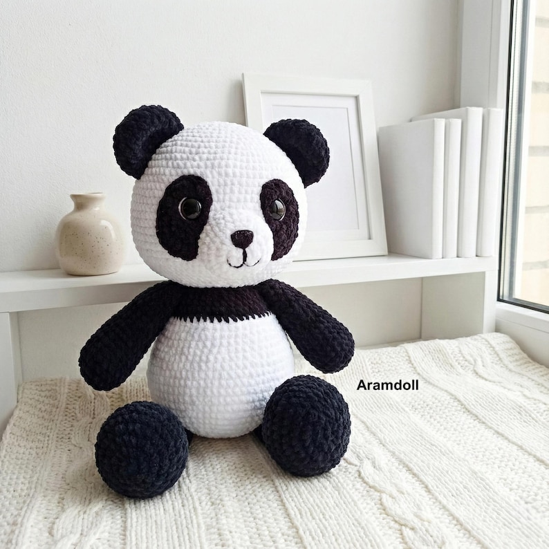 May include: A handmade crochet panda bear doll with a white body, black arms, legs, and ears. The panda has black eye patches and a small, embroidered smile. The doll is sitting on a white surface.