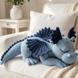 5 ALT TEXT FOR PHOTOS
1) Crochet sleeping dragon plush with pale blue wings resting on a white sofa in a bright neutral room
2) Close-up of the crocheted dragon head with embroidered sleepy eyes and soft textured stitches
3) Side view of amigurumi dragon showing sculpted wings, horns, and back spikes in pastel tones
4) Crochet dragon plush posed on a cozy knit blanket with minimalist nursery-style decor in the background
