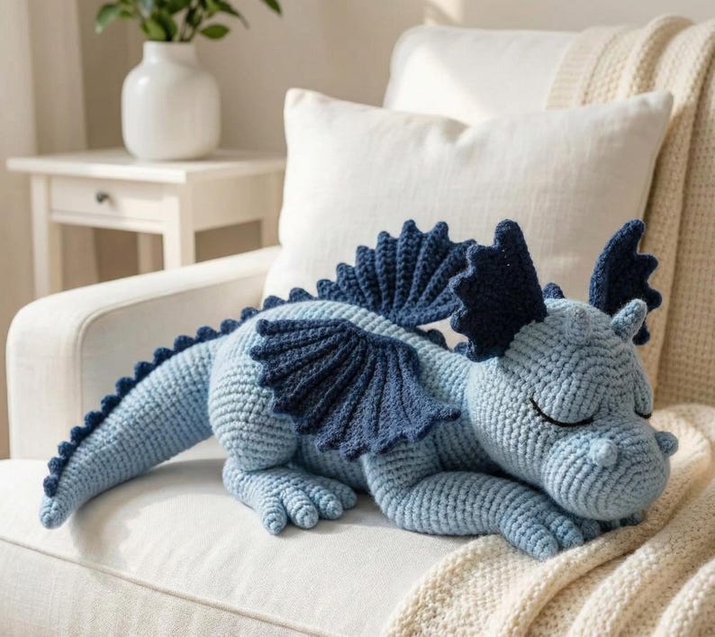 5 ALT TEXT FOR PHOTOS
1) Crochet sleeping dragon plush with pale blue wings resting on a white sofa in a bright neutral room
2) Close-up of the crocheted dragon head with embroidered sleepy eyes and soft textured stitches
3) Side view of amigurumi dragon showing sculpted wings, horns, and back spikes in pastel tones
4) Crochet dragon plush posed on a cozy knit blanket with minimalist nursery-style decor in the background