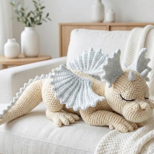 5 ALT TEXT FOR PHOTOS
1) Crochet sleeping dragon plush with pale blue wings resting on a white sofa in a bright neutral room
2) Close-up of the crocheted dragon head with embroidered sleepy eyes and soft textured stitches
3) Side view of amigurumi dragon showing sculpted wings, horns, and back spikes in pastel tones
4) Crochet dragon plush posed on a cozy knit blanket with minimalist nursery-style decor in the background