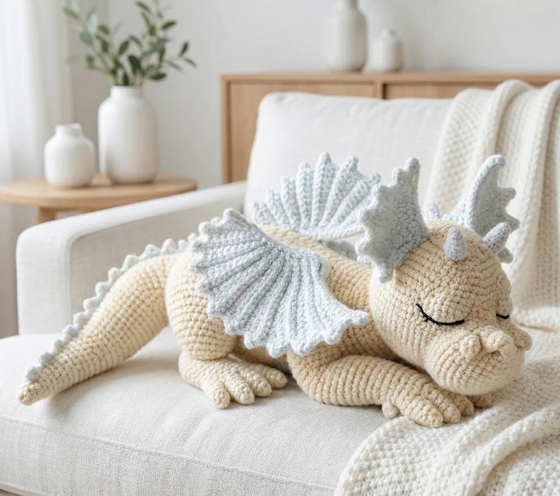 5 ALT TEXT FOR PHOTOS
1) Crochet sleeping dragon plush with pale blue wings resting on a white sofa in a bright neutral room
2) Close-up of the crocheted dragon head with embroidered sleepy eyes and soft textured stitches
3) Side view of amigurumi dragon showing sculpted wings, horns, and back spikes in pastel tones
4) Crochet dragon plush posed on a cozy knit blanket with minimalist nursery-style decor in the background