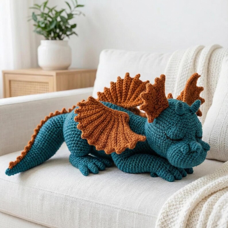May include: A handmade crochet dragon in teal and brown, resting on a white sofa. The dragon features detailed wings and a long tail. The soft sculpture is a whimsical decorative item.