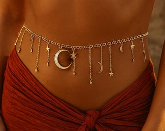 May include: A gold-colored body chain featuring a celestial design. The chain is adorned with dangling charms of stars, moons, and small crystals. The main focal point is a large crescent moon pendant. The chain is worn over a red fabric.