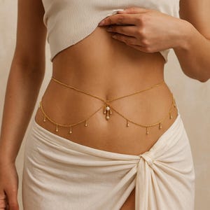 Puede incluir: Una delicada cadena de cuerpo dorada. La cadena presenta un diseño de doble hebra que cae sobre la cintura. Pequeños dijes colgantes y un colgante central de perlas añaden detalles. La joya se usa sobre una falda blanca.