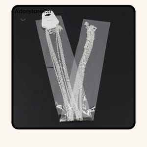May include: Two silver necklaces in clear plastic packaging. One necklace has multiple strands, and the other has a strand of small white beads. The packaging has a white tag with the text "AdorstoreCo".