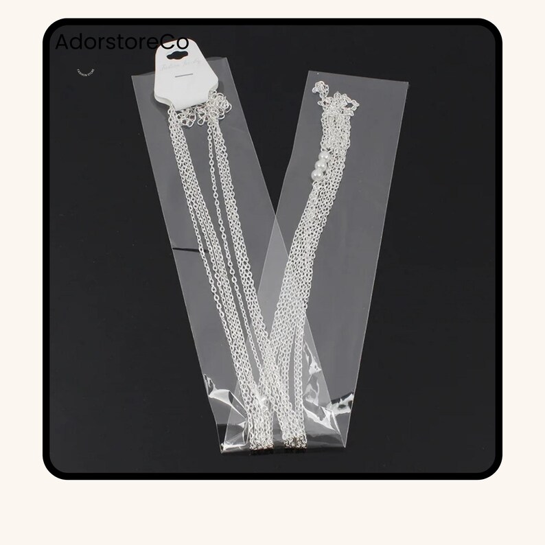 May include: Two silver necklaces in clear plastic packaging. One necklace has multiple strands, and the other has a strand of small white beads. The packaging has a white tag with the text "AdorstoreCo".