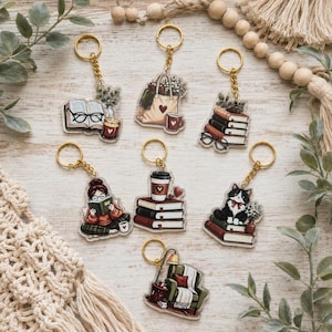 May include: A collection of seven acrylic keychains featuring illustrated designs of books, a reading person, a cat, a tote bag, a coffee cup, and an armchair. Each keychain has a gold-toned metal ring and displays detailed, colorful artwork.