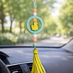 May include: A car charm featuring a yellow rubber duck inside a light green ring. The charm is adorned with pastel-colored beads and a yellow tassel. It hangs from a car's rearview mirror, adding a touch of whimsy to the vehicle's interior.