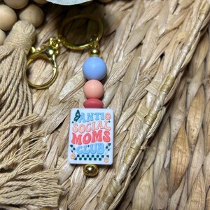 May include: A gold-coloured keyring with a rectangular charm reading "ANTI SOCIAL MOMS CLUB" in a retro font. The keyring has blue, pink, and red beads. The background is a woven texture.