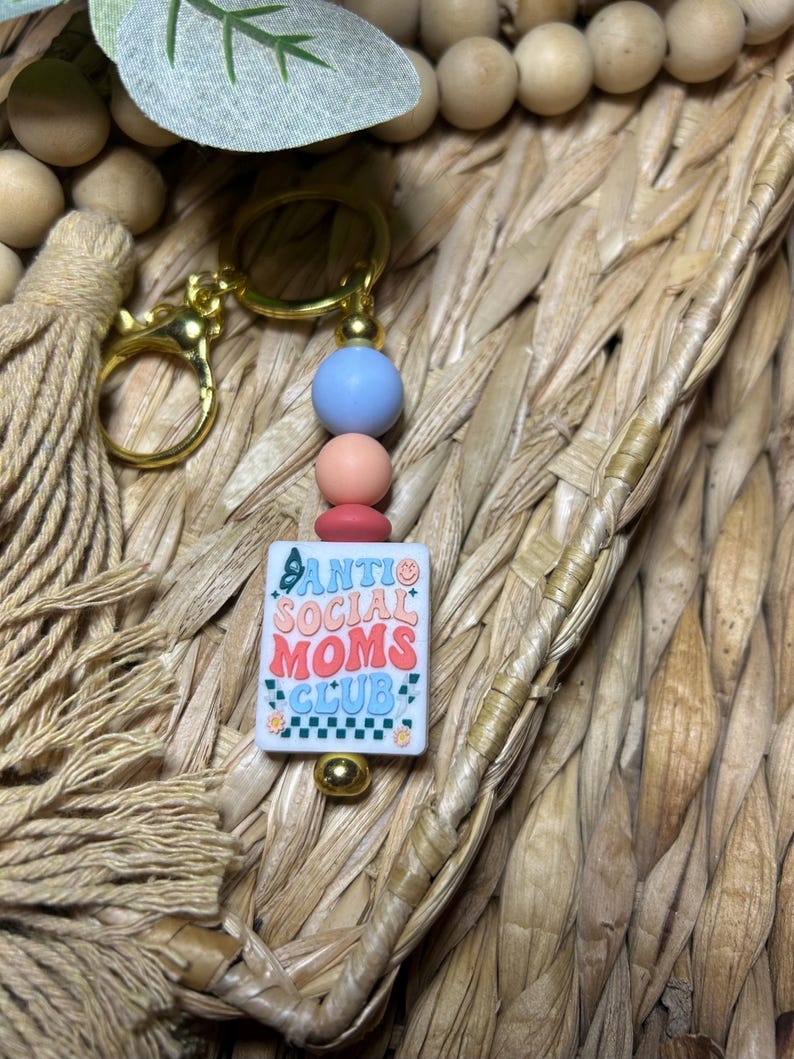 May include: A gold-coloured keyring with a rectangular charm reading "ANTI SOCIAL MOMS CLUB" in a retro font. The keyring has blue, pink, and red beads. The background is a woven texture.