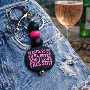 May include: A black keychain with the text "IT COSTS $0.00 TO BE PETTY AND I LOVE FREE SHIT" in pink. The keychain has black and pink beads and a silver clasp. The keychain is attached to a pair of distressed denim shorts.