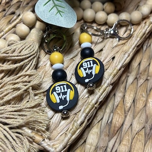 May include: Two keychains with black, yellow, and white beads. Each keychain features a black circular charm with the number "911" and a sound wave graphic inside a yellow headphone design. The keychains are on a woven background.