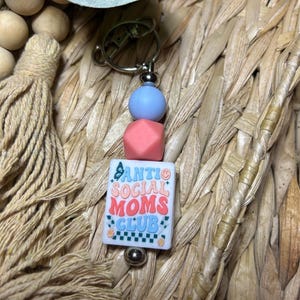 May include: A keychain with a silver clasp, blue and coral beads, and a white square charm. The charm has the text "Anti Social Moms Club" in a retro font with butterfly and flower accents. The background is a woven texture.