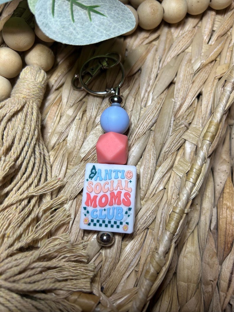 May include: A keychain with a silver clasp, blue and coral beads, and a white square charm. The charm has the text "Anti Social Moms Club" in a retro font with butterfly and flower accents. The background is a woven texture.