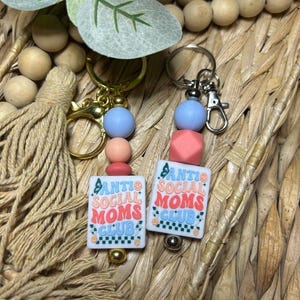 May include: Two keychains with the text "Anti Social Moms Club" on white rectangular beads. Each keychain has a gold or silver clasp, with blue, pink, and peach-coloured beads. The keychains are set against a woven background.