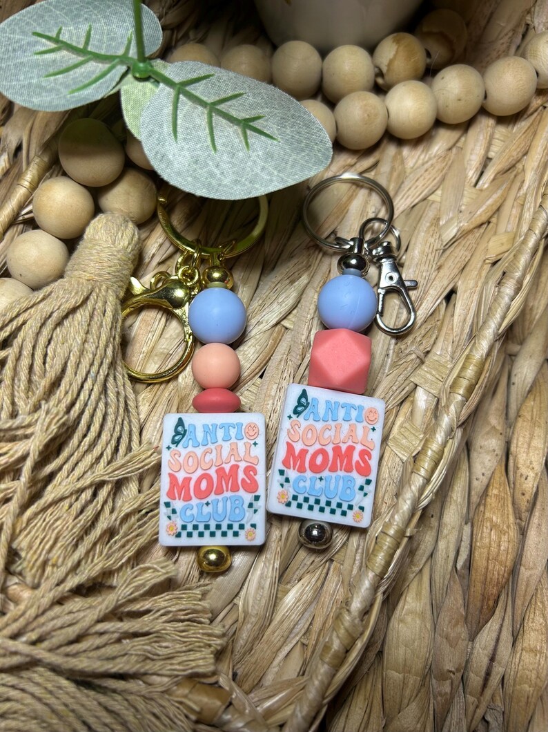 May include: Two keychains with the text "Anti Social Moms Club" on white rectangular beads. Each keychain has a gold or silver clasp, with blue, pink, and peach-coloured beads. The keychains are set against a woven background.