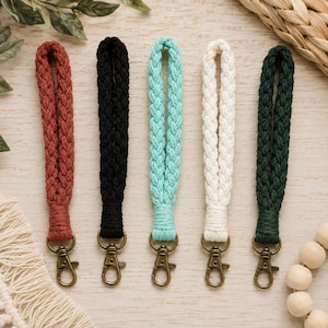 May include: Five braided macrame wristlet lanyards in rust, black, turquoise, cream, and dark green. Each lanyard features a woven cord loop secured with a wrapped knot and finished with a bronze-toned metal lobster clasp for attaching keys.