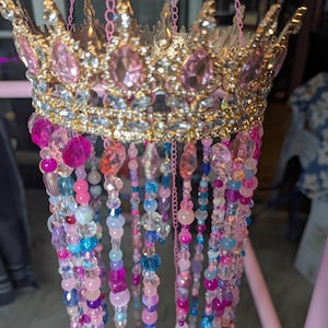 May include: A gold-toned crown adorned with pink and clear faceted crystals, featuring multiple hanging strands of multi-colored beads in shades of pink, blue, and purple. The crown is suspended by thin pink chains attached to the metal frame.