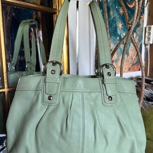 Könnte beinhalten: Eine salbeigrüne Handtasche aus Leder mit zwei Schulterriemen mit silberfarbenen Metallschnallen, einer plissierten Vorderseite und dem eingeprägten Markennamen Coach in der Mitte. Die Tasche hat eine strukturierte Form mit sichtbaren Nähten.
