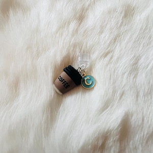 Puede incluir: Un dije de taza de café en miniatura con la palabra "COFFEE" impresa en el lateral. El dije tiene una tapa negra y se combina con un dije circular turquesa y dorado con la letra "C".