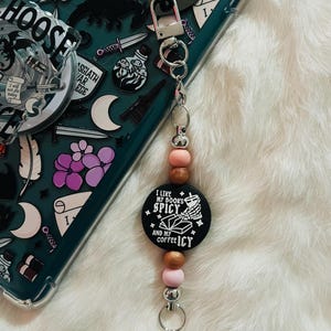 May include: A keychain with a black circular charm that reads "I like my books spicy and my coffee icy." The charm is accented with pink and brown beads and a silver clasp. The background is a phone case with a dark teal design.
