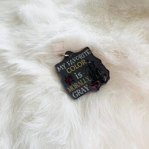 Puede incluir: Una insignia negra con el texto "MY FAVORITE COLOR is MORALLY GRAY" en amarillo y blanco. La insignia tiene un clip de plástico transparente y un diseño de silueta y rosas rojas.
