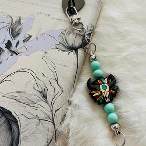 May include: A keychain with a black and turquoise bull skull charm, turquoise beads, and silver accents. The keychain is attached to a floral patterned wallet. The background is a white, fluffy surface.