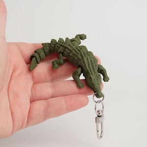 Peut inclure: Un porte-clés en forme d'alligator vert avec un fermoir argenté. L'alligator est imprimé en 3D avec des détails texturés, notamment des écailles et des pattes et une queue articulées. Le porte-clés est tenu dans une main sur fond blanc.
