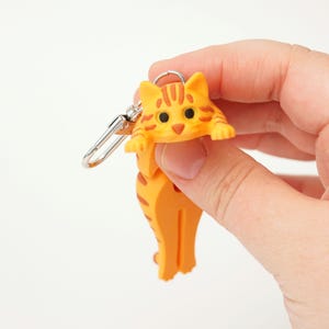 Puede incluir: Llavero de gato naranja y marrón con cierre plateado. El gato tiene un patrón de rayas y una pose juguetona. El llavero se sostiene sobre un fondo blanco.