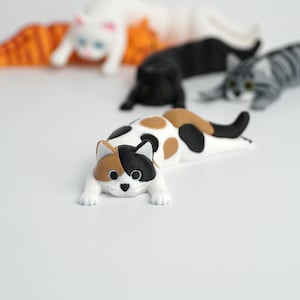 Op de afbeelding: Een verzameling kattenbeeldjes in verschillende kleuren, waaronder wit, zwart, grijs en lapjeskat. De katten liggen ontspannen, met gedetailleerde kenmerken. De beeldjes zijn waarschijnlijk decoratieve items of speelgoed.