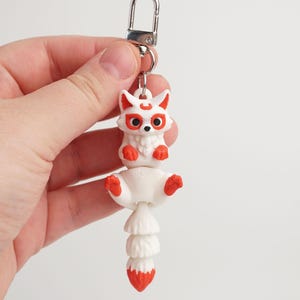 May include: A white and red fox keyring with a silver clasp. The fox has red accents on its ears, eyes, and feet. The tail is layered and ends in a red tip. The keyring is held by a hand.