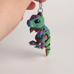 Könnte beinhalten: Ein farbenfroher, beweglicher Dinosaurier-Schlüsselanhänger. Die kleine, 3D-gedruckte Figur hat einen Farbverlauf aus Blau-, Grün- und Lilatönen. Er hat einen silberfarbenen Verschluss zum Befestigen an Schlüsseln oder Taschen. Der Dinosaurier ist ein lustiges, einzigartiges Accessoire.