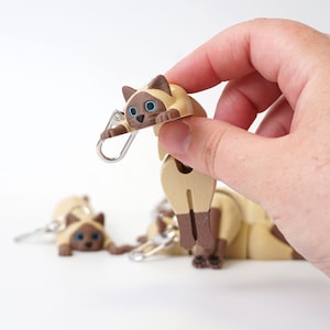Può includere: Portachiavi a forma di gatto siamese marrone e beige con occhi blu e chiusura argentata. Diversi portachiavi simili sono visibili sullo sfondo. I portachiavi a forma di gatto sono realizzati in materiale liscio e presentano dettagli.