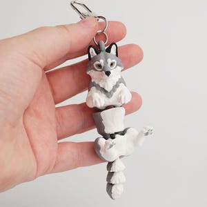 Könnte beinhalten: Ein grau-weißer Wolf-Schlüsselanhänger mit einem silbernen Verschluss. Der Wolf ist segmentiert, wobei die obere Hälfte aufrecht und die untere Hälfte in einer spielerischen Pose ist. Der Schlüsselanhänger wird in einer Hand vor weißem Hintergrund gehalten.