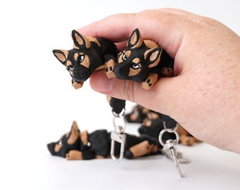 May include: Black and tan miniature dog figurines, some attached to keychains. The small dog sculptures are lying down and have detailed features. The figurines are held in a hand against a white background.