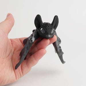 Può includere: Piccola statuetta di pipistrello nero con grandi orecchie e grandi occhi. Il pipistrello ha le ali spiegate ed è tenuto in una mano su sfondo bianco. La statuetta ha una superficie strutturata.