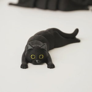 Puede incluir: Figura de gato negro con ojos amarillos, tumbada con las patas extendidas. El gato está hecho de un material texturizado y está colocado sobre una superficie blanca. Otras figuras de gato similares son visibles en el fondo.